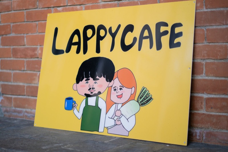 レンガの壁に立てかけた、LAPPY CAFEの黄色い看板。宿の2人のイラストが描かれています