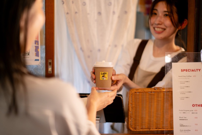 LAPPY CAFEのカウンターで、佳鈴が紙カップのコーヒーを手渡しているところ
