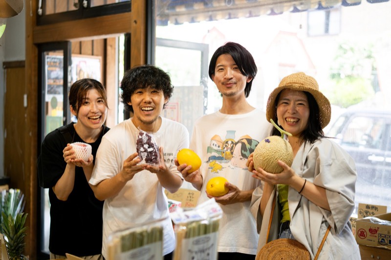 八百屋さんで、メロンや果物を手に笑う4人